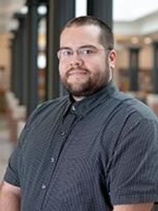 A man with short hair, glasses, and a beard wearing a dark checkered shirt stands indoors with blurred columns and windows.