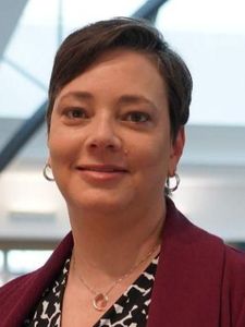 A person with short dark hair wearing a red sweater looks directly at the camera against a blurred background.