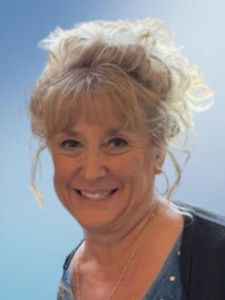 A smiling woman with curly blonde hair pulled up and wearing a blue top and dark cardigan stands against a light blue.
