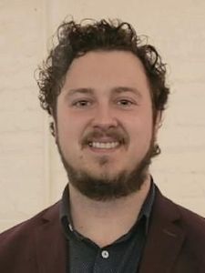 A man with curly dark hair and a beard smiles while wearing a dark blazer and shirt against a light brick wall.