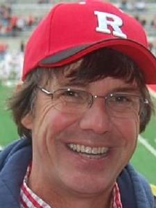 A man wearing glasses and a red baseball cap with a white letter R smiles at a sports stadium with players on the field.