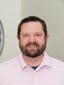 A man with short dark hair and a beard wearing a light purple button-up shirt stands indoors near black columns.