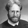 A black and white portrait of a man with glasses, a beard, and curly hair wearing a sweater over a collared shirt.