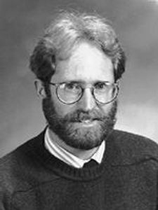 A black and white portrait of a man with glasses, a beard, and curly hair wearing a sweater over a collared shirt.