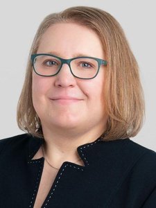 A person with light brown hair and teal glasses smiles slightly while wearing a black jacket against a plain gray background.