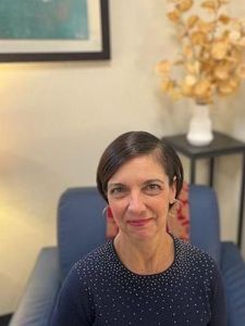A woman with short dark hair and a navy blue top with small white dots sits on a blue chair in a softly lit room.