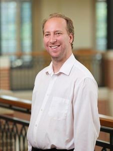A man with light brown hair wearing a white button-up shirt smiles while standing indoors near a railing.
