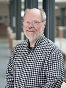 A smiling middle-aged man with glasses and a beard wearing a black and white checkered shirt stands indoors.