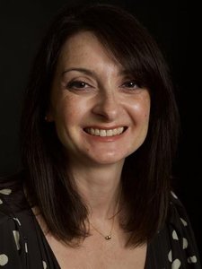 A woman with straight dark brown hair smiles gently against a black background, wearing a black top with white spots.