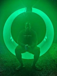 A person sits barefoot inside a large glowing green circular structure at night, holding a can.