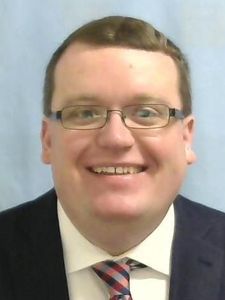 A man with short brown hair and glasses smiles wearing a dark suit, white shirt, and striped tie against a light blue.