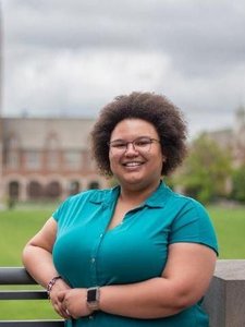 A smiling person with curly hair and glasses stands outdoors wearing a teal shirt, with a building and green lawn behind.