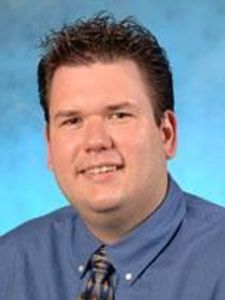 A man with short dark hair wearing a blue shirt and patterned tie smiles against a blue gradient background.