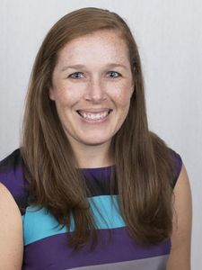 A woman with long brown hair and freckles smiles at the camera wearing a purple and blue striped sleeveless top.