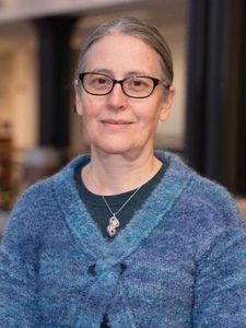 A person with glasses and gray hair tied back smiles slightly while leaning on a wooden railing indoors.