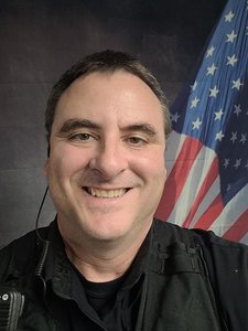 A man wearing a black shirt and vest smiles at the camera with an American flag in the background.