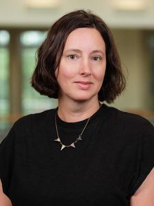 A woman with short brown hair wearing a black shirt and a triangular pendant necklace looks directly at the camera.