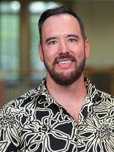A man with short dark hair and a beard smiles while wearing a black and white floral shirt indoors.