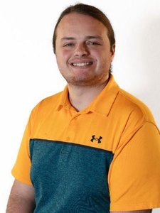 A JCU faculty member smiles warmly for a portrait, wearing an orange and dark teal polo shirt.