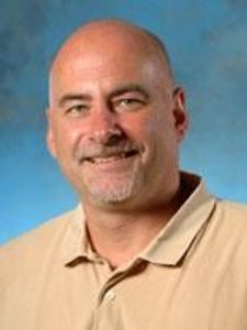 A middle-aged man with a shaved head and goatee smiles slightly while wearing a beige collared shirt against a blue.