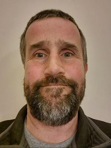A man with short dark hair and a full beard looks directly at the camera against a plain background.