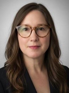 A woman with long brown hair and glasses wearing a blue button-up shirt stands indoors near a railing.