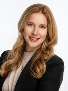 A woman with long wavy blonde hair smiles wearing a black blazer and light-colored top against a white background.