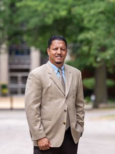 A man in a beige blazer, blue shirt, patterned tie, and dark pants stands on a street with trees and a building behind him.