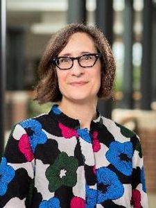 A person with short brown hair and glasses wearing a colorful floral-patterned shirt stands indoors with columns and brick.