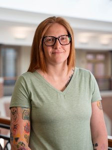 A person with straight red hair, black glasses, and a green shirt stands indoors showing colorful tattoos on their arms.