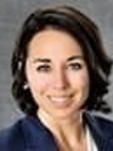 A JCU faculty member smiles warmly, wearing a dark blazer. Her short, dark hair frames her face against a gray background.