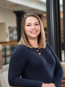 A woman with shoulder-length blonde hair smiles while leaning on a wooden railing inside a building.