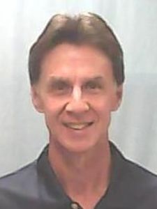 A man with short brown hair smiles slightly while wearing a dark collared shirt against a plain light background.