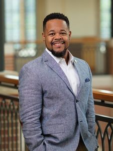 A man in a light blue blazer and white shirt smiles while standing indoors near a railing with blurred windows behind him.