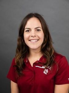 A woman with long brown hair smiles while wearing a maroon polo shirt against a gray background.