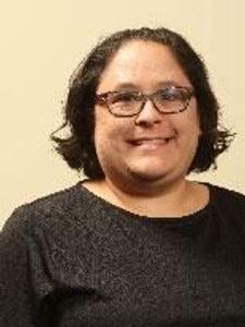 A person with shoulder-length dark hair and glasses smiles while wearing a dark sweater against a plain beige background.