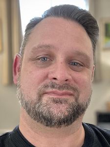 A man with short dark hair and a trimmed beard looks into the camera wearing a black shirt with wooden buttons.