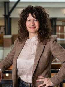 A woman with curly dark hair smiles while leaning on a railing inside a building with brick and metal accents.