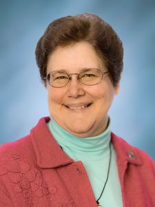 A smiling woman with short brown hair and glasses wears a pink jacket over a light blue turtleneck against a blue background.
