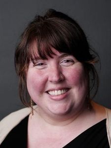 A woman with long brown hair and a nose ring smiles gently against a dark teal background.