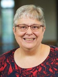 A smiling older woman with short gray hair and glasses wearing a patterned top.