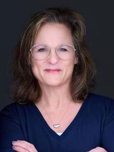 A woman with shoulder-length brown hair and glasses smiles gently against a dark background.