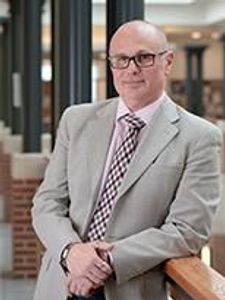 A bald man wearing glasses and a light gray suit stands indoors, leaning on a wooden railing and looking at the camera.