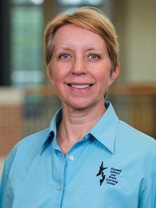 A woman with short blonde hair smiles wearing a light blue shirt with a library and learning center logo.