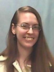 A woman with long brown hair and glasses smiles slightly against a plain light blue background.