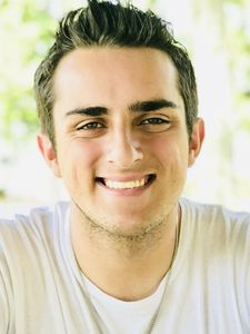 A young man with short dark hair smiles directly at the camera wearing a white shirt and a silver necklace.