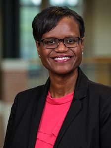 A woman with short black hair and glasses smiles while wearing a black blazer over a bright pink top indoors.