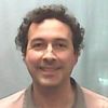 A man with curly dark hair and a light brown shirt smiles gently against a plain light gray background.