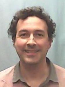 A man with curly dark hair and a light brown shirt smiles gently against a plain light gray background.