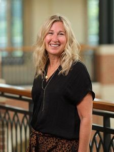A smiling woman with wavy blonde hair wearing a black blouse and leopard print pants stands indoors by a railing.